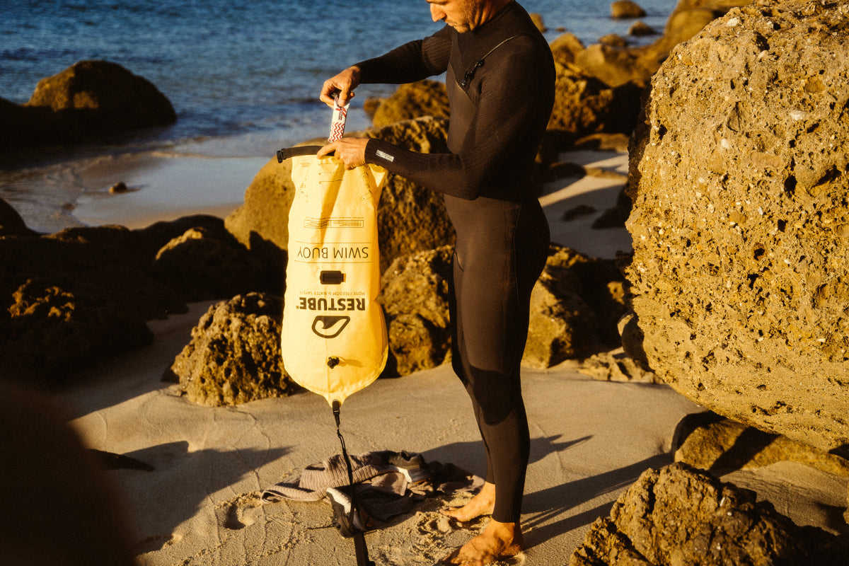 Se muestra a un nadador guardando alimentacion deportiva en la seccion de bolsa seca de la boya de senalizacion RESTUBE PRO, preparandose para entrar a nadar en el mar
