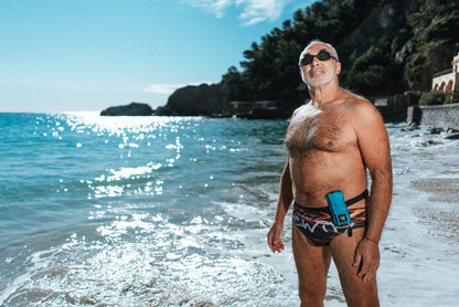 Se muestra a un hombre portando un dispositivo RESTUBE ACTIVE de color azul en su cintura al costado izquierdo. El hombre esta por entrar a nadar equipado con RESTUBE, para mayor seguridad en los entrenamientos de natación en aguas abiertas